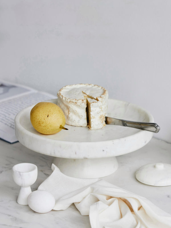 Marble cake stand with a piece of cake and a lemon on a light gray surface.