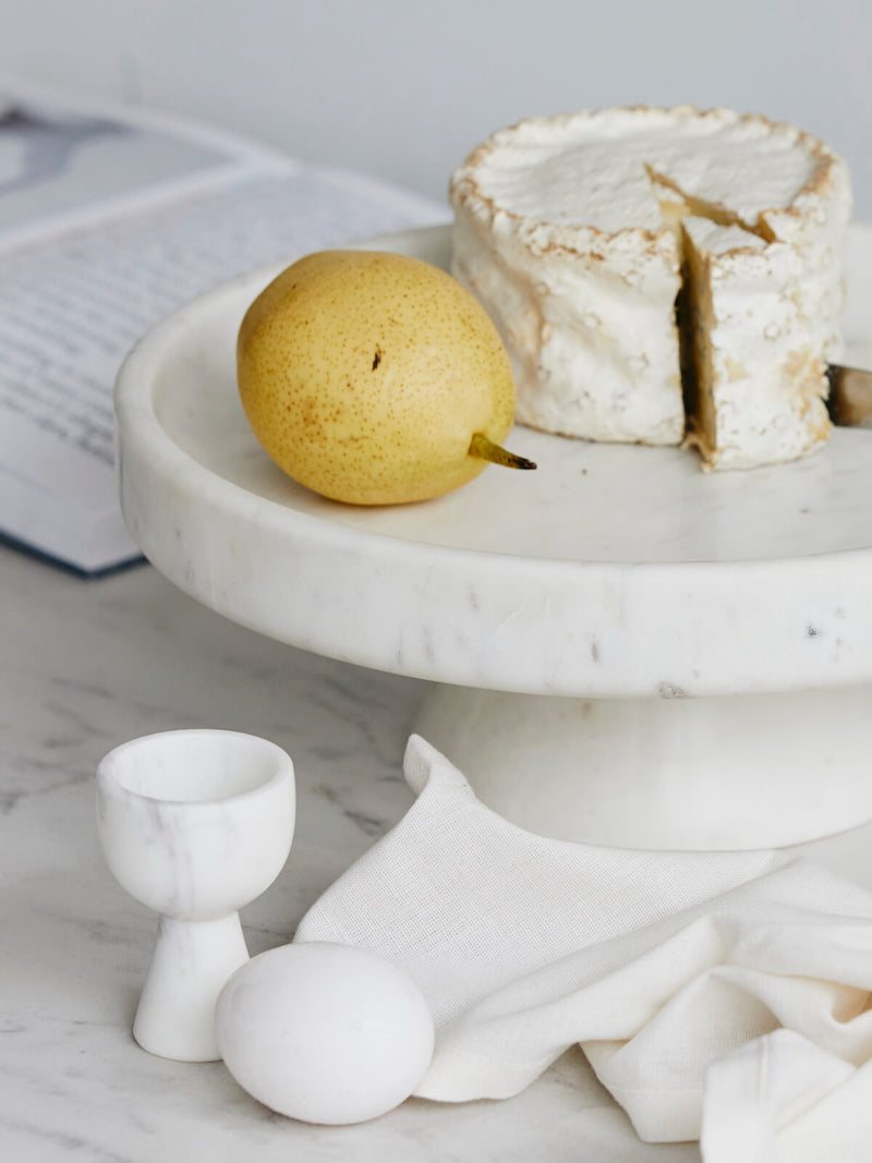 Marble cake stand with a pear, wedge of cheese, and white ceramic items on a marble surface.