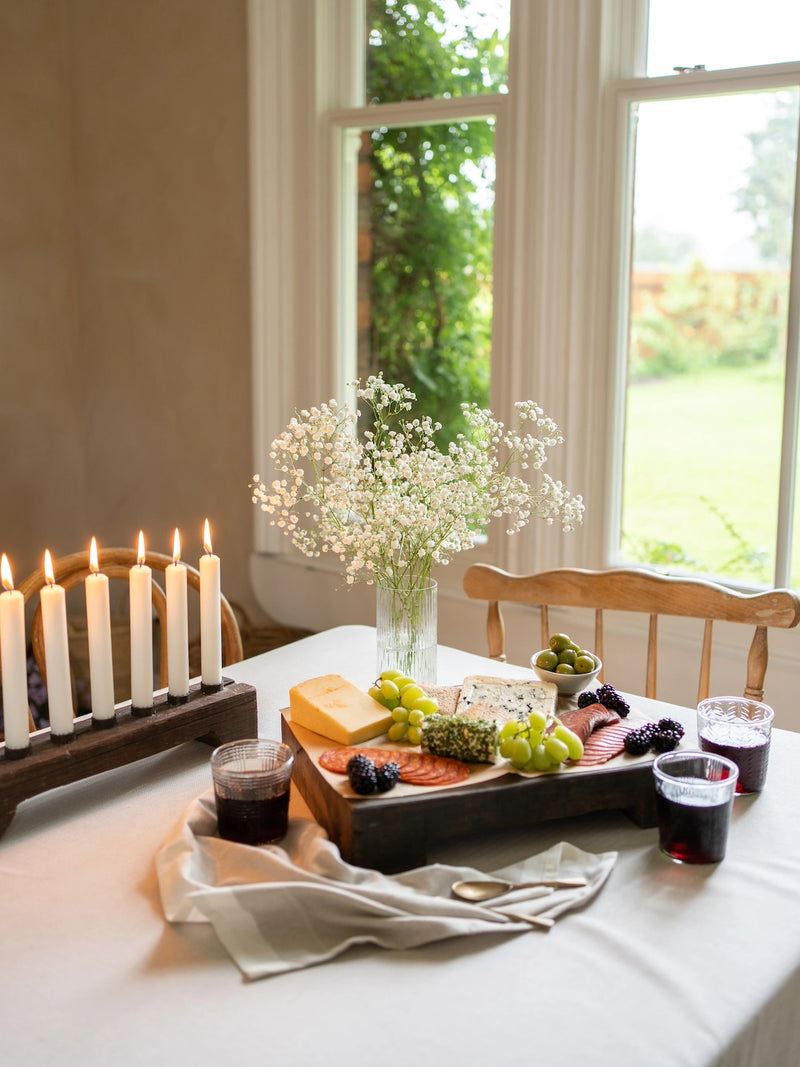 Dining table with a cheeseboard patla tray, fruits, and wooden candle holder with white candles in a bright room.