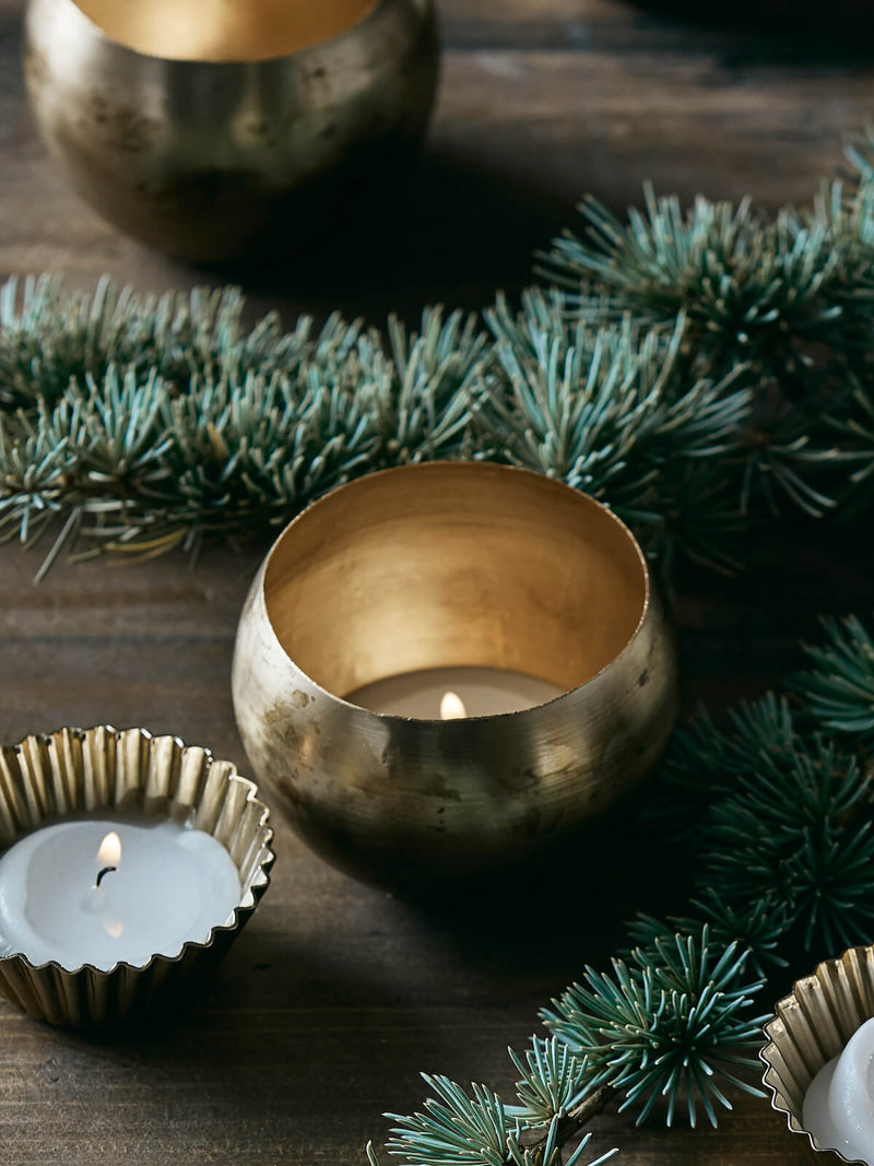 Brass tealight holder with candles lit in a festive setting, greenery on top of rustic table.