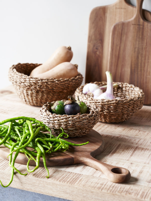 This image shows a collection of woven seagrass baskets, some filled with vegetables, on a wooden surface. The baskets vary in size, with the smallest holding green peppers and a purple vegetable, possibly an eggplant. Larger baskets contain garlic, butternut squash, and a pile of long green beans or peppers. Wooden cutting boards are also visible in the background, adding to the natural and rustic aesthetic of the scene.