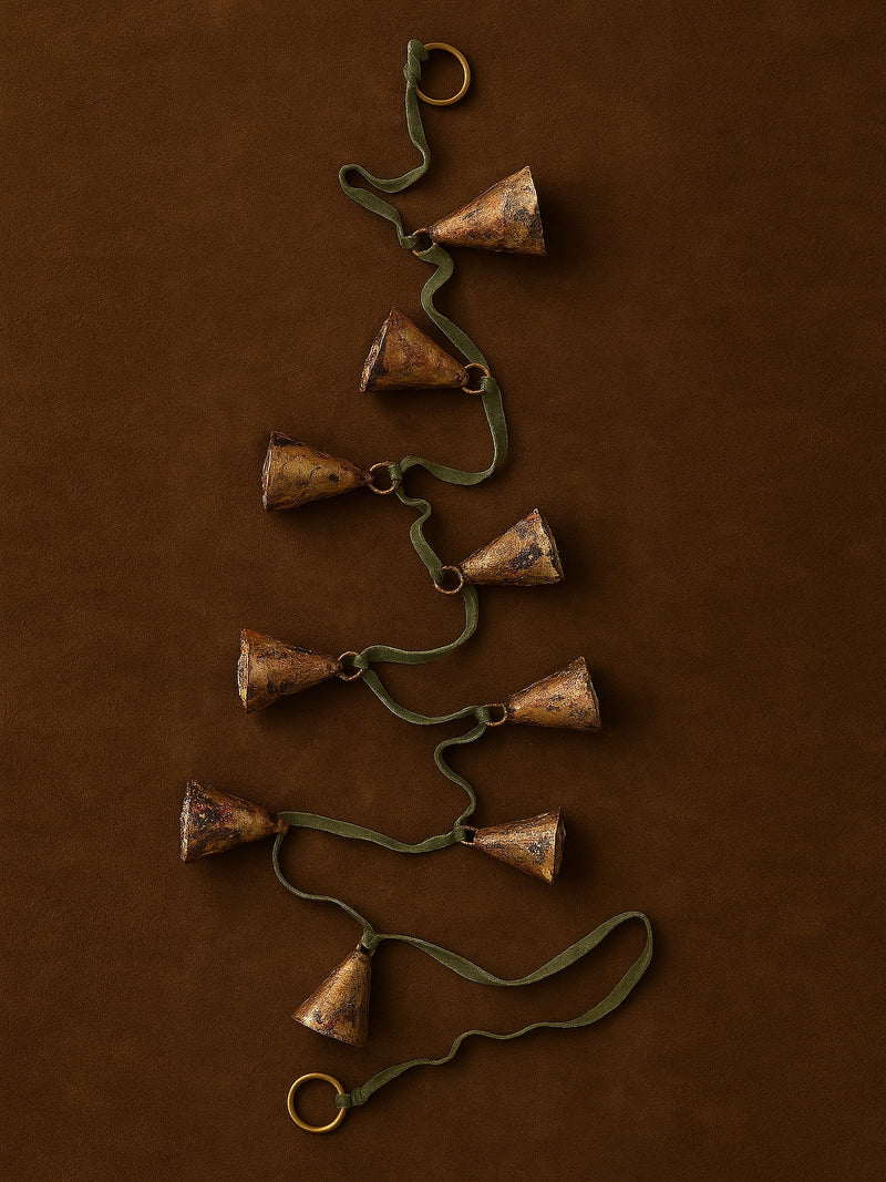 Decorative brass bells garland on a green ribbon string by THATCH Living against a brown background.