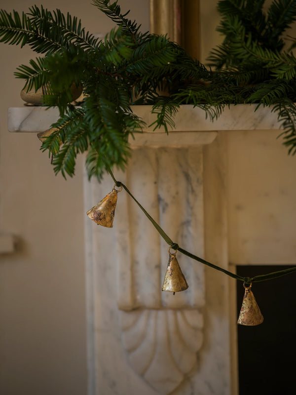 Decorative brass bells garland on a green ribbon string by THATCH Living against a marble fireplace.