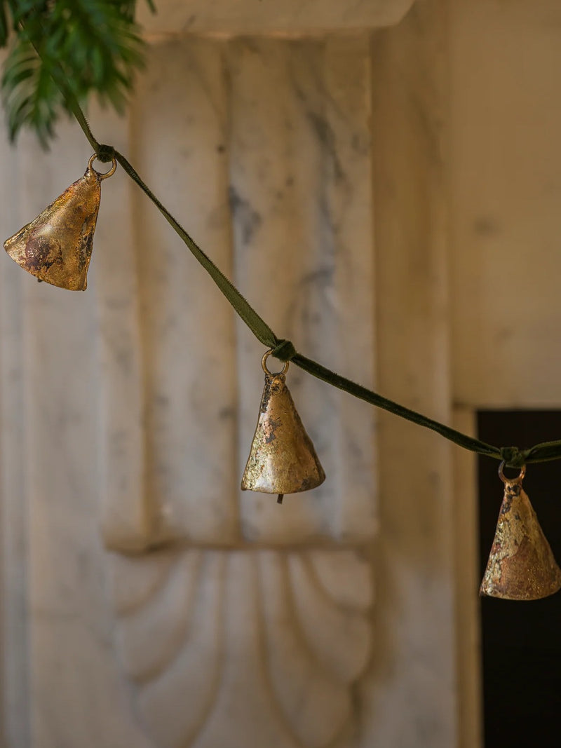 Decorative brass bells garland on a green ribbon string by THATCH Living against a marble fireplace.