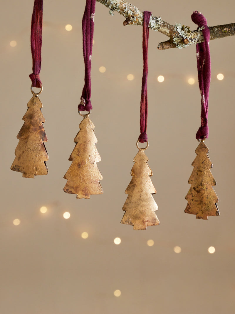 Four gold tree-shaped ornaments by THATCH Living hanging from a branch against a beige background with soft lights.