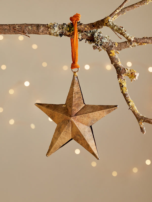 Gold decorative metal distressed star by THATCH Living hanging from a branch with a blurred light background.
