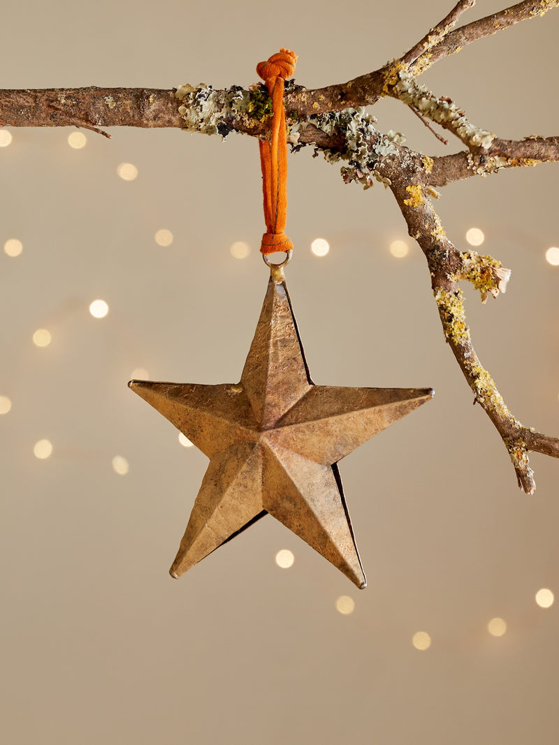 Gold decorative metal distressed star by THATCH Living hanging from a branch with a blurred light background.