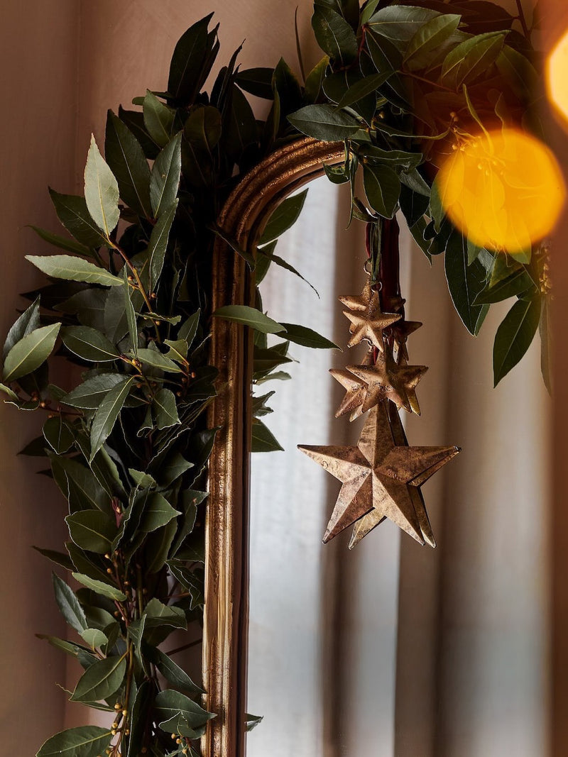 Decorative wreath with green leaves and star-shaped hanging gold ornaments by THATCH Living on a mirror.