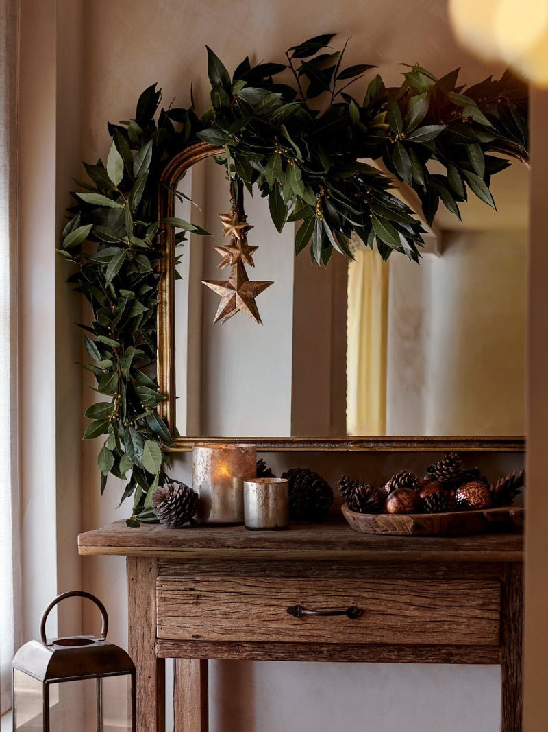 Decorative setup with a mirror, hanging gold stars by THATCH Living, greenery, and candles on a wooden surface.