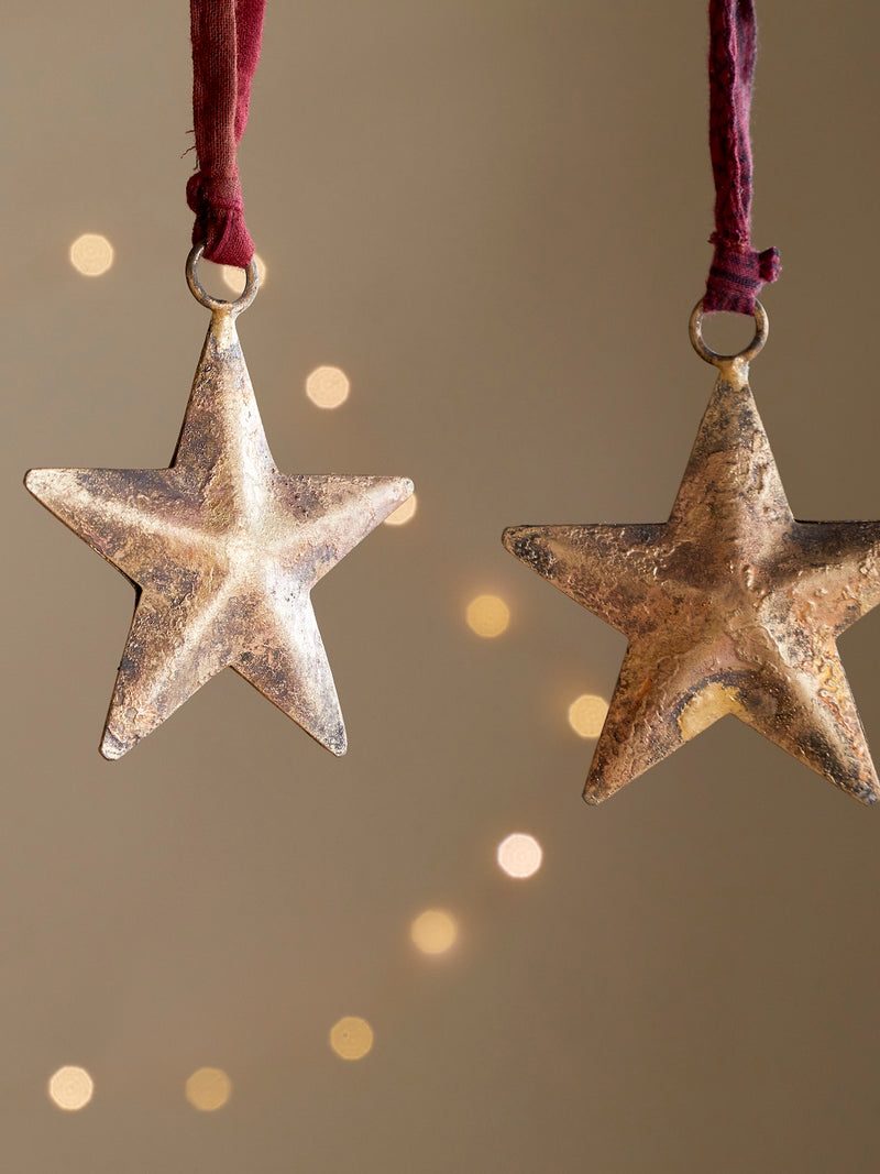 Two star-shaped metal distressed rustic ornaments by THATCH Living hanging against a soft, blurred background with bokeh lights.