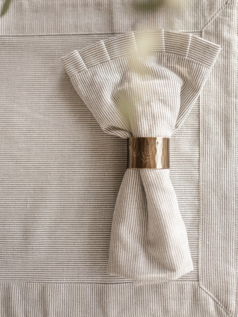Folded white napkin with a gold napkin ring on a textured beige surface by THATCH Living