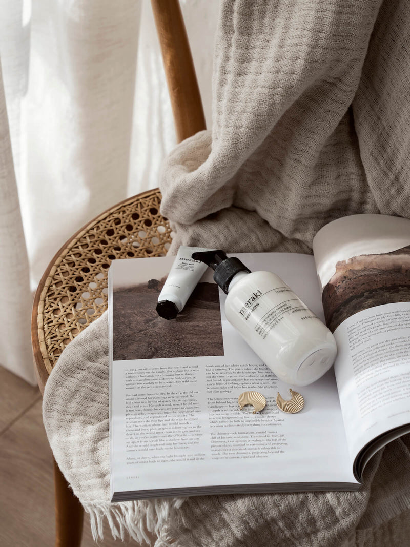 A bottle of Meraki Silky Mist hand lotion on top of chair, surrounded by magazine, earings in a calm slow moving environment. 