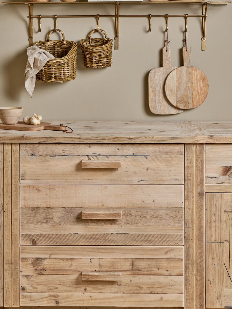 Wooden kitchen counter with drawers, cutting boards, and baskets on a neutral wall. Wall hung rattan baskets by THATCH Living.